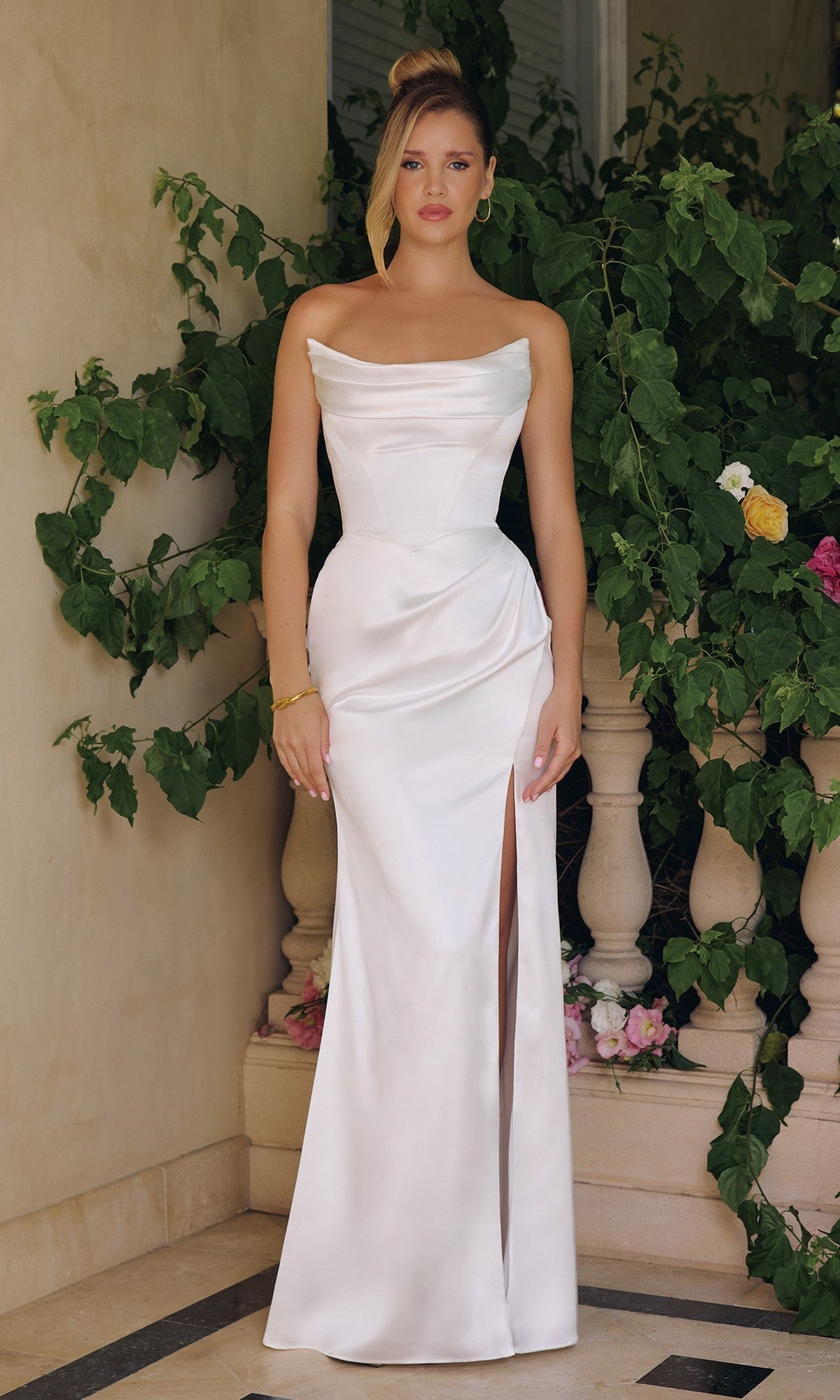 A woman stands before greenery, wearing the Amoris T820W long prom dress—white satin with off-the-shoulder straps, a floor-length skirt, and a daring leg slit.